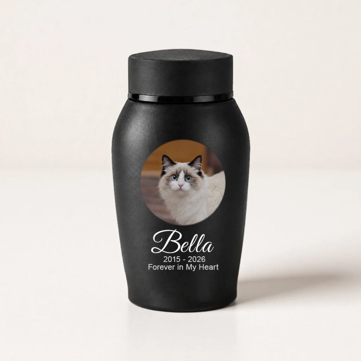A matte black pet memorial urn with a lid, featuring a printed photo of a white and brown cat with the text 'Bella 2015 - 2026 Forever in My Heart' on a light beige background.