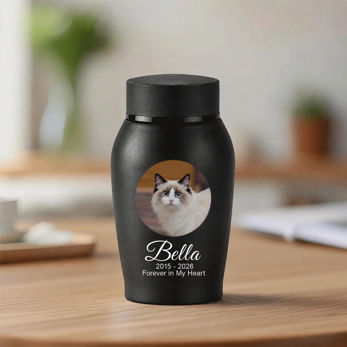 A matte black pet memorial urn with a custom photo of a cat and the text 'Bella 2015 - 2026 Forever in My Heart' on a wooden table in a blurred indoor setting.