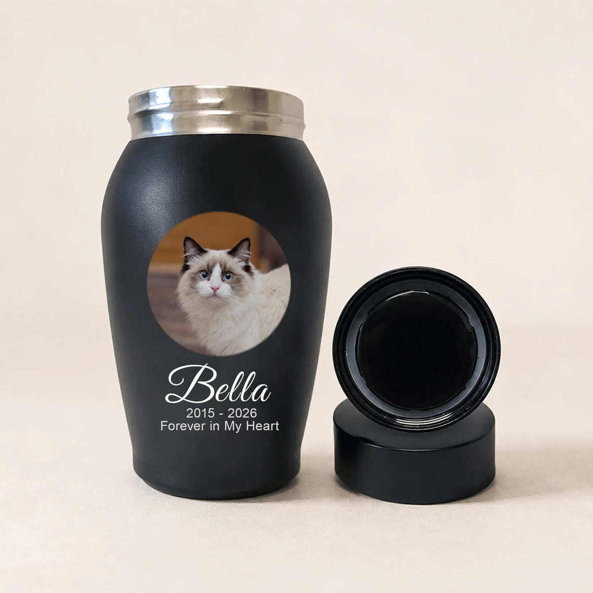 A matte black pet memorial tumbler with a silver lid, shown next to its detached black lid on a light beige background. The tumbler features a circular photo of a white and brown cat with the text 'Bella 2015 - 2026 Forever in My Heart' printed below.