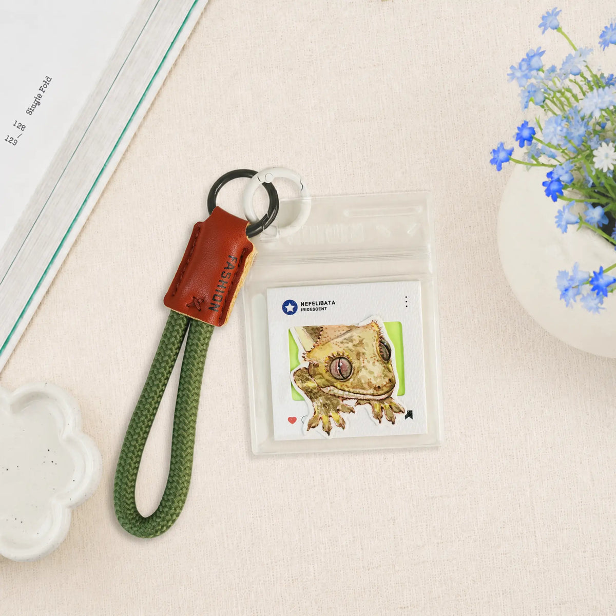 A clear plastic card holder keychain with a green braided strap, a brown leather tab, and a black metal ring, shown on a light beige textured surface next to a book, a small dish, and blue flowers.