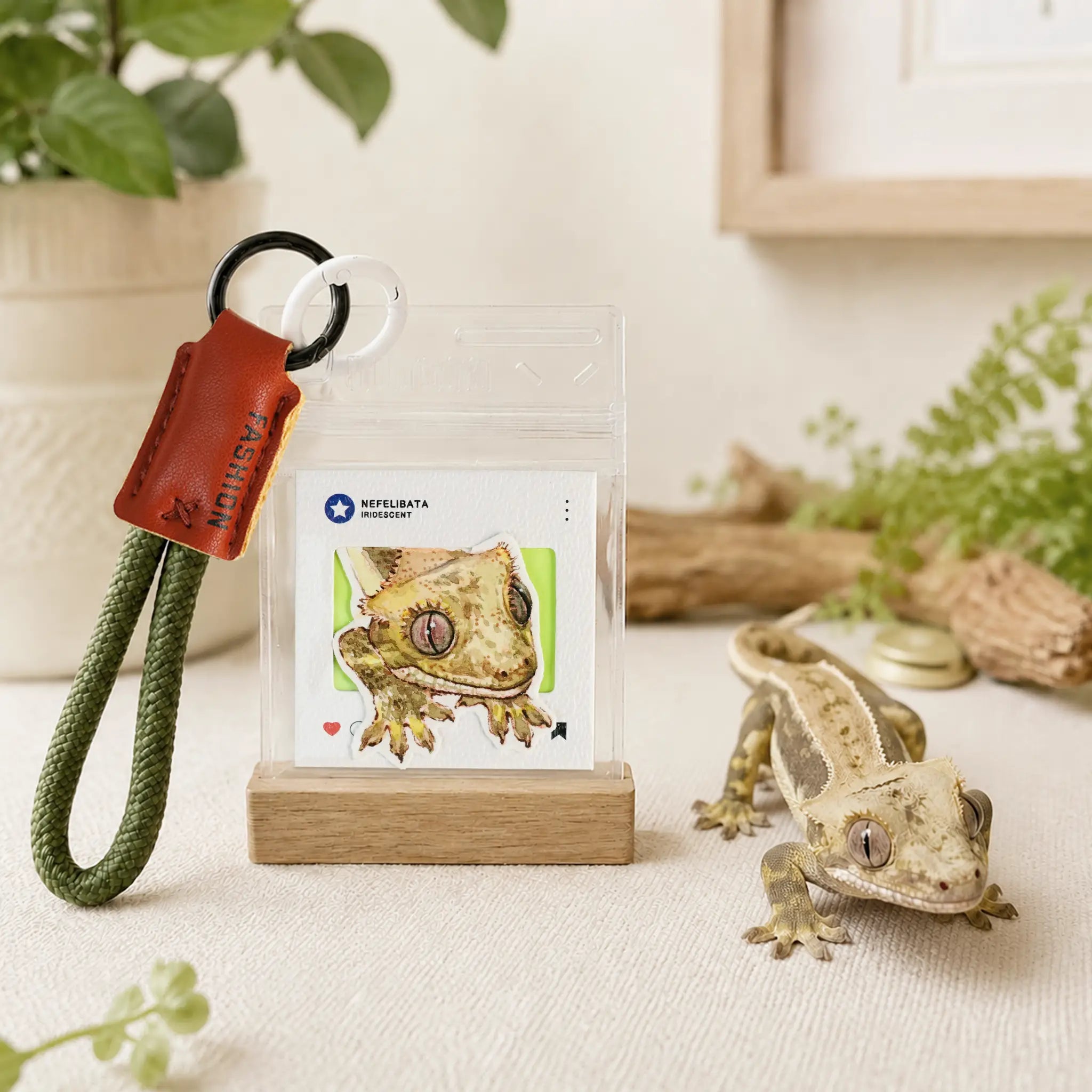 A small gecko keychain accessory displayed on a clear stand with a wooden base, alongside a real gecko figurine and soft home decor props. The keychain includes a green braided cord, a black ring, and an orange-brown leather tag with the brand name visible.