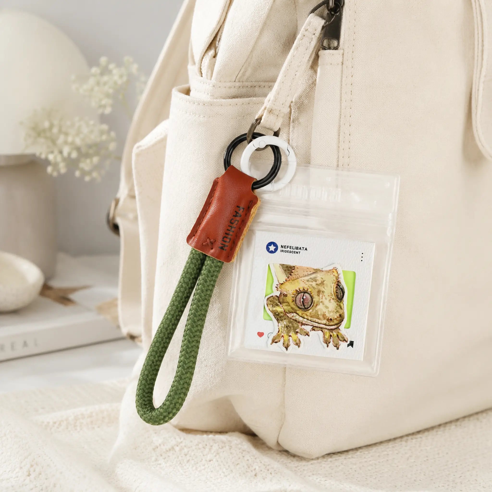A close-up of a cream canvas backpack with a hanging clear ID badge holder and keychain. The holder contains a card with a chameleon illustration and the text 'Chameleon' and 'Chameleon Park'.
