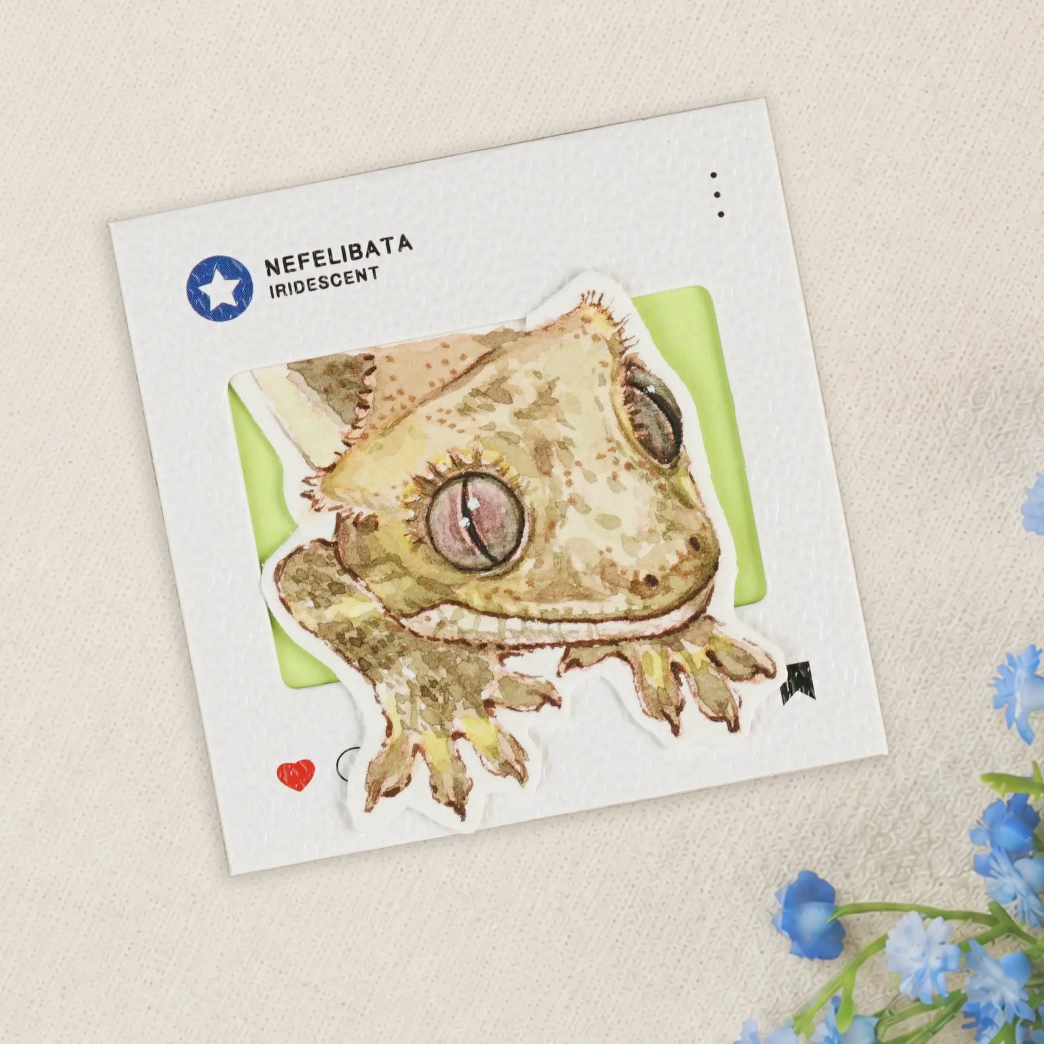A die-cut sticker of a green-toad-like amphibian with iridescent beige and brown coloring, displayed on a white backing card labeled 'NEFELIBATA IRIDESCENT' against a light beige textured background with small blue flowers in the corner.